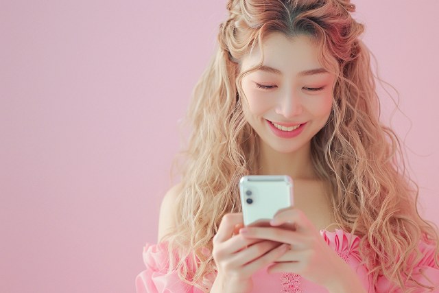 ピンクの壁の前でスマホを持っているピンクのドレス姿のキャバ嬢