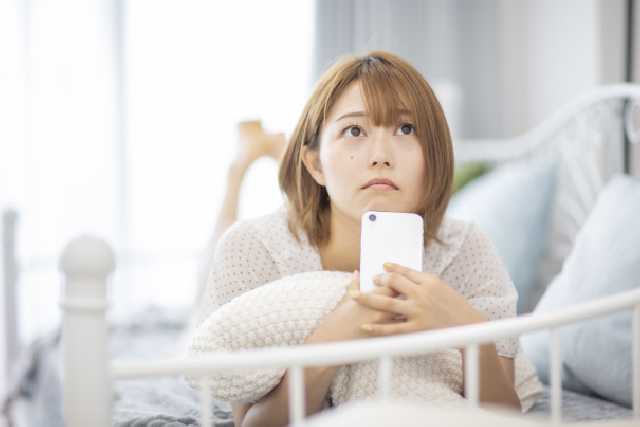 スマホを手に持った女性がベットにうつぶせになって何かを思い浮かべる様子