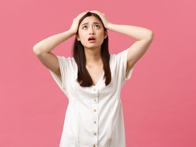 ピンク色の壁の前で頭亜を抱えてたつ若い女性