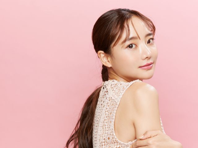 ピンク色の壁の前に横向きに立ち振り向くようにこちらを見る若い日本人女性