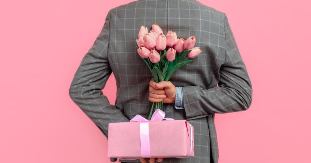 ピンクの背景の前で花束とプレゼントを背中に隠している男性