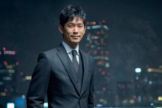夜景を背景に微笑んで立つスーツ姿の日本人男性