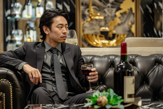 黒い革張りのソファに座ってワインを飲むスーツ姿の日本人の男性