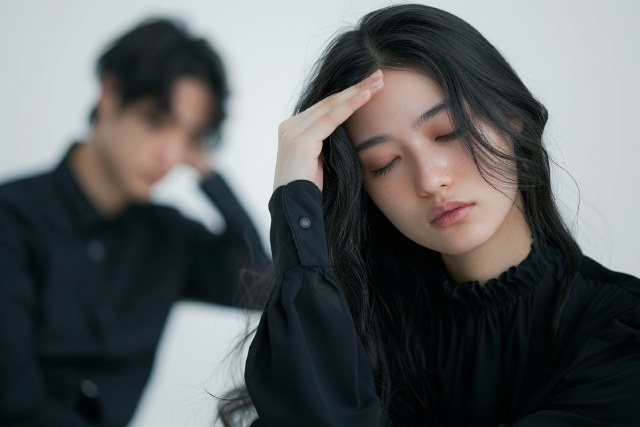 額に手を当て、苦しそうな表情で頭を抱える日本人の若いロングヘアの女性と背後にいる男性