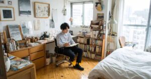 日本人男性が寝室のデスクに置かれたオフィスチェアに座り、ビジネスカジュアルな服装で読書をしながらコーヒーを飲んでいる