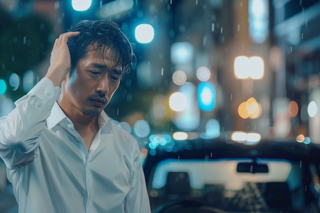 雨が降る夜の街でつらそうに頭を抱える白シャツの日本人男性