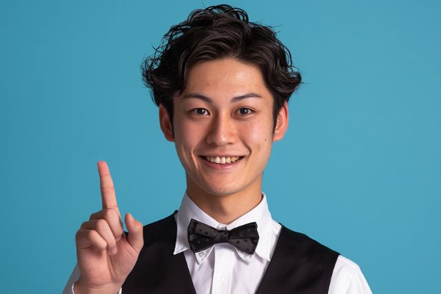 人差し指を上に向けて笑顔を見せる黒い蝶ネクタイとベストを着用している男性