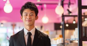 ピンク色のコンセプトカフェを背景に笑顔で立つ黒いスーツ姿の男性