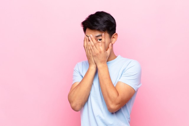 手で顔を隠しつつ覗き見をする白いTシャツの男性