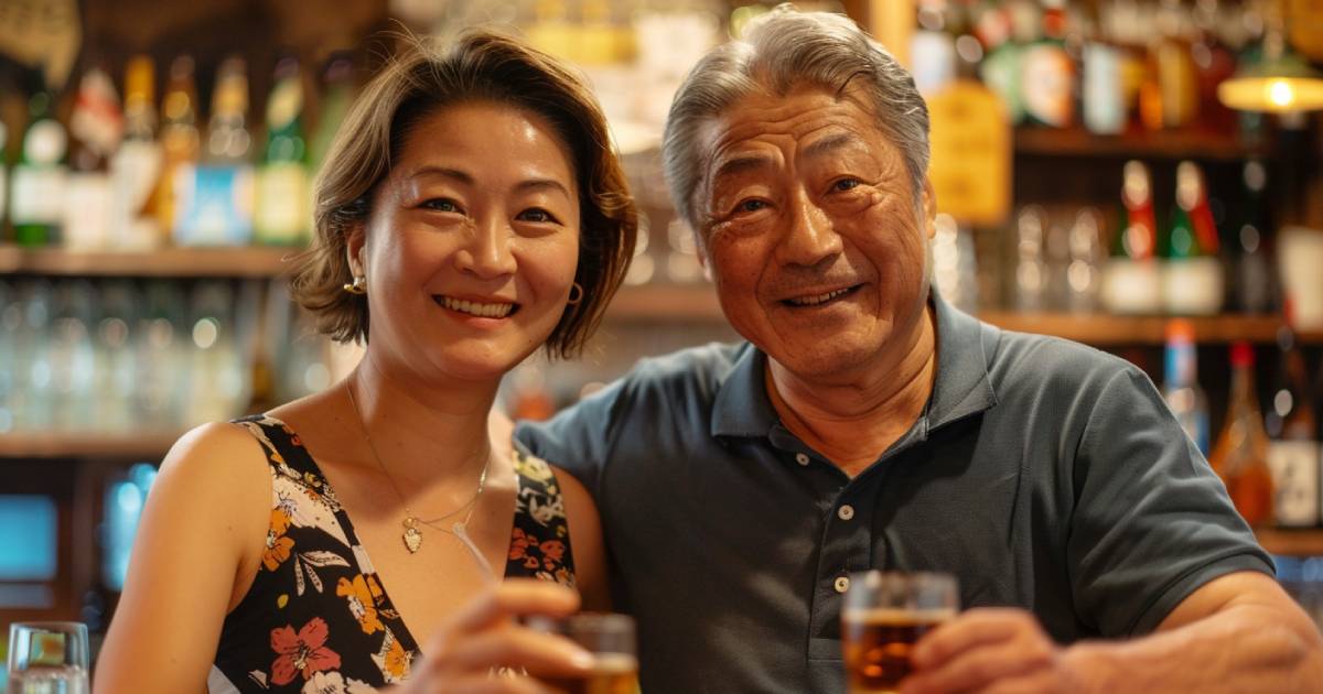 ポロシャツを着た中年の日本人男性と花柄のワンピースを着た女性が、ショットグラスを手にカメラに向かって微笑んでいる
