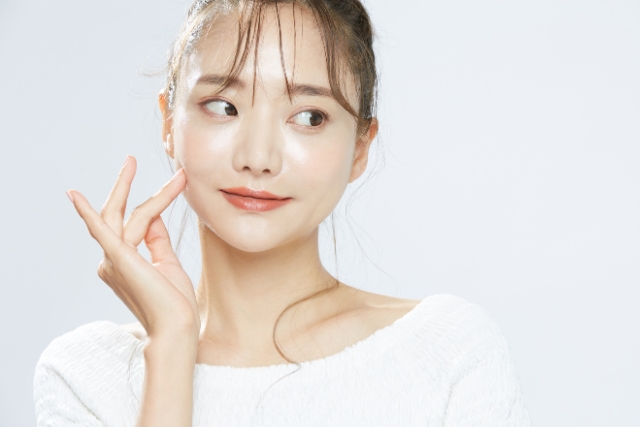 白い壁の前で白いトップスを着て頬に指を当てる若い女性