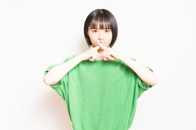 緑のトップスを着て口の前でばつ印を作る黒髪ボブの若い女性