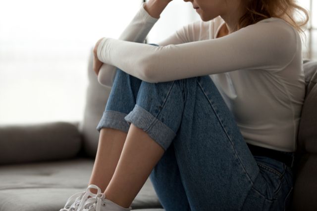 ソファに体育座りで頭を抱えるデニム姿の女性